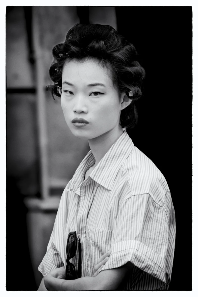 Portrait en noir et blanc d’une personne avec une coiffure rétro et un regard direct, vêtue d’une chemise à fines rayures