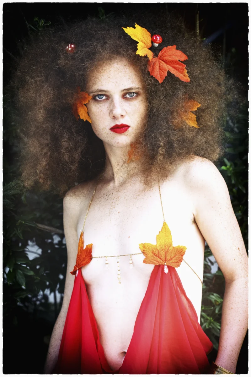 Personnage vêtu d’une robe rouge avec des feuilles d’automne dans les cheveux et sur une parure, pose devant un feuillage
