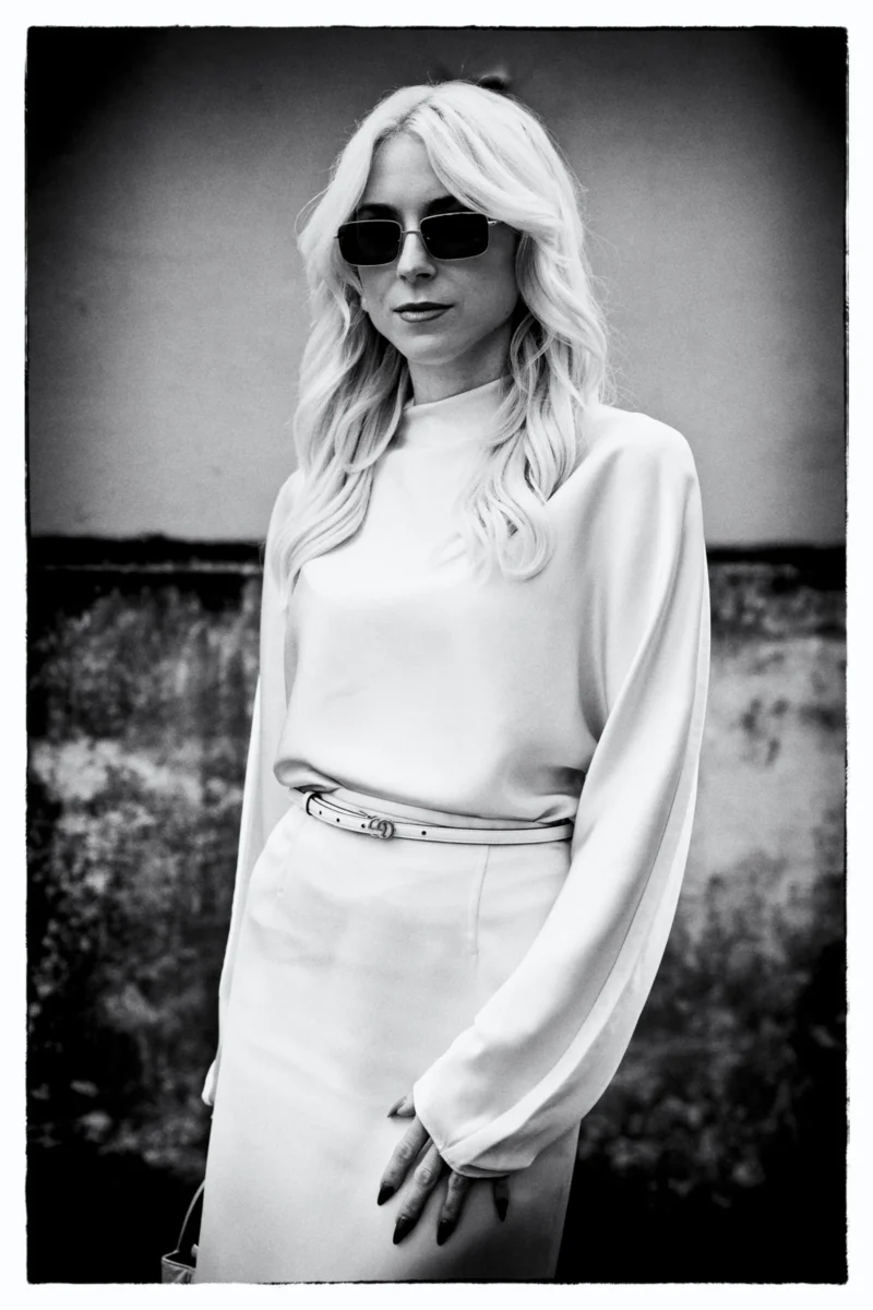 Portrait en noir et blanc d’une personne aux cheveux longs portant des lunettes de soleil et une tenue élégante cintrée