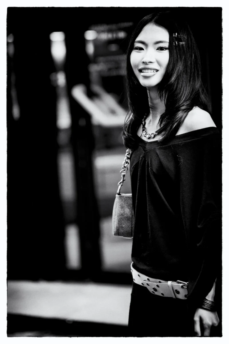 Portrait en noir et blanc d’une personne souriante portant une tenue élégante avec un sac à main, en extérieur devant