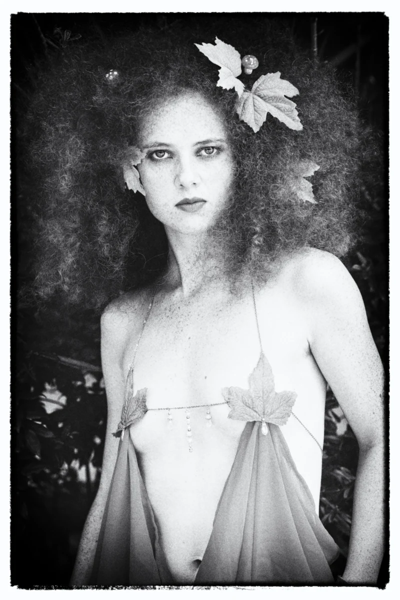 Portrait en noir et blanc d’une personne vêtue d’une tenue légère ornée de feuilles, cheveux volumineux et regard intense