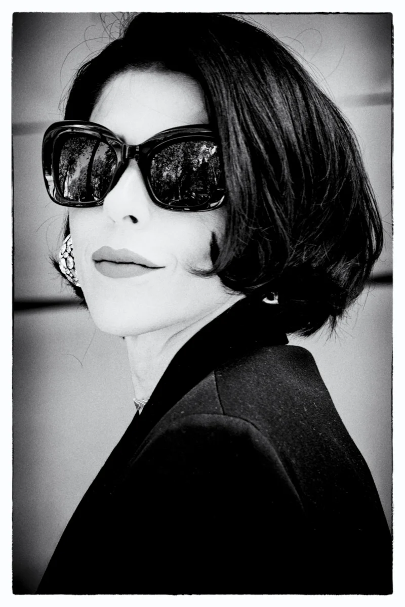 Portrait en noir et blanc d'une personne portant des lunettes de soleil cat-eye et une veste élégante, sourire discret.
