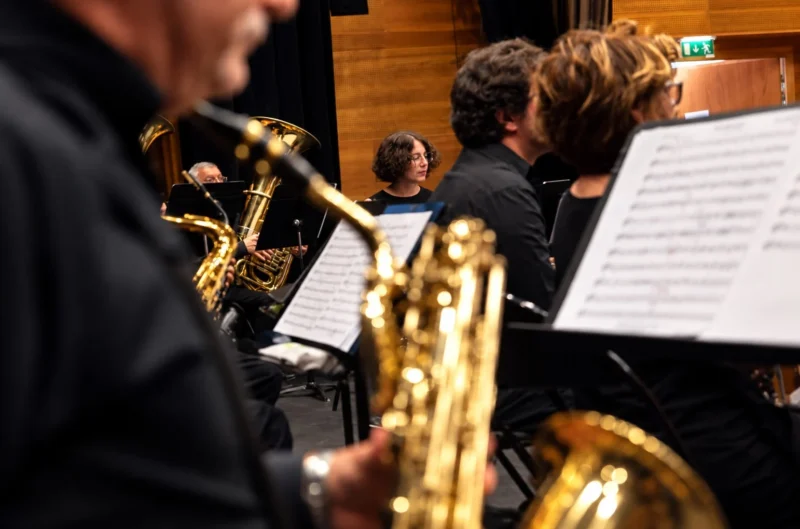 Musiciens jouant des instruments à vent dans un orchestre, avec des partitions devant eux, ambiance concert en intérieur.