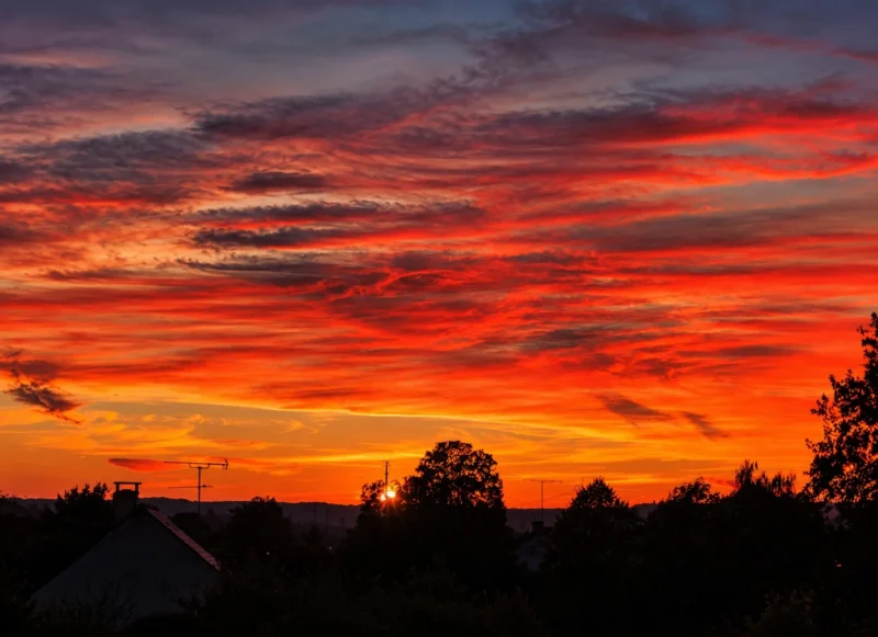Couchant aux teintes orangées et rouges sur un horizon avec des nuages et des silhouettes d'arbres et de maisons en contrebas