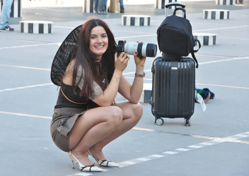 Une personne accroupie tenant un appareil photo avec un grand objectif, souriante, près d'un bagage à roulettes et d'un sac