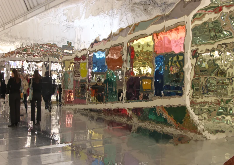 Installation artistique murale colorée dans une galerie, reflétée sur un sol brillant, avec des visiteurs en arrière-plan.