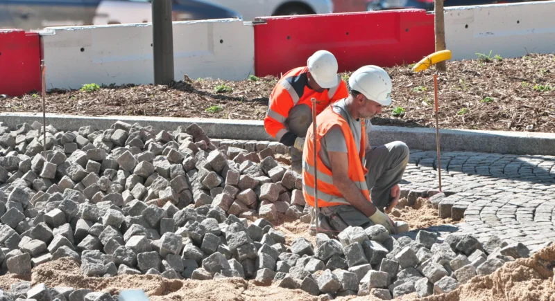 Deux travailleurs en tenue de sécurité manipulant des pierres concassées sur un chantier de construction près d’un trottoir.