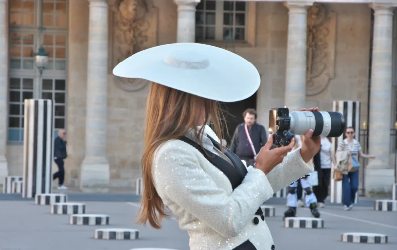 Une personne en tenue élégante avec un chapeau large prend une photo avec un appareil photo professionnel devant un bâtiment