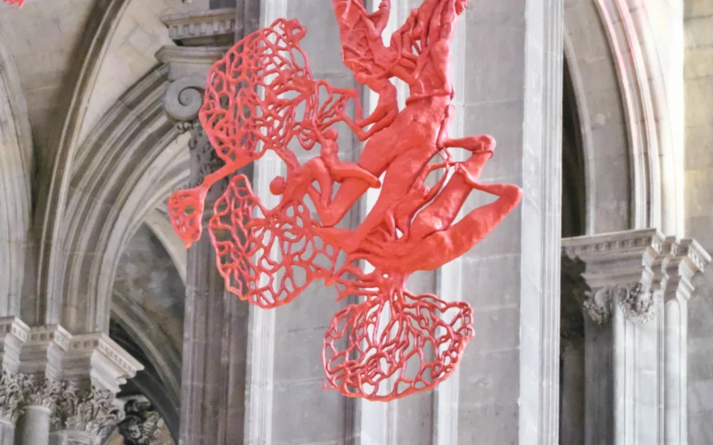 Jean paul · 05/10/2026 · 2400 x 1498 Sculpture rouge en forme de structure organique suspendue dans un cadre architectural gothique en pierre grise.
