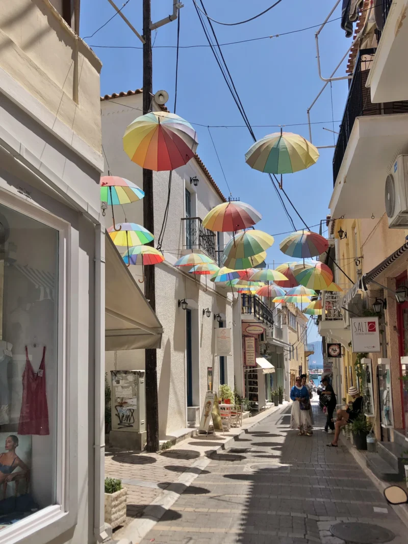 Jean paul · 21/06/2024 · 1800 x 2400 Rue étroite décorée d'ombrelles colorées suspendues entre les bâtiments, ambiance chaleureuse et commerçante sous un ciel