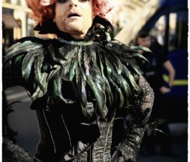 Thierry · 03/10/2026 · 1600 x 2400 Personne déguisée en costume extravagant avec une perruque rose et des plumes vertes sur une rue animée, expression