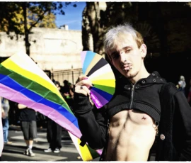 Thierry · 03/10/2026 · 2400 x 1600 Un individu tenant des drapeaux arc-en-ciel et un éventail coloré lors d'une manifestation en extérieur sous un ciel dégagé.