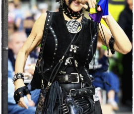 Thierry · 03/10/2026 · 1600 x 2400 Personne vêtue d’un costume gothique et punk mangeant un sandwich dans une rue animée, avec accessoires et maquillage