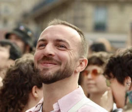 Francoise · 03/10/2026 · 2400 x 1600 Un homme souriant au milieu d'une foule en extérieur, entouré de personnes, avec un fond architectural flou.