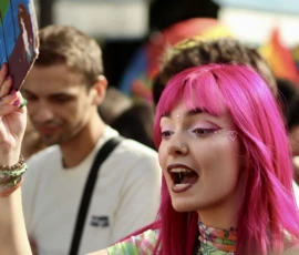Francoise · 03/10/2026 · 2400 x 2400 Personne aux cheveux roses vêtue de couleurs vives, souriante et tenant un accessoire coloré, lors d'un événement festif
