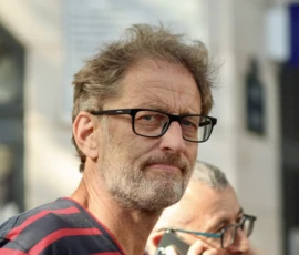 Francoise · 03/10/2026 · 2400 x 1600 Portrait d’un homme en extérieur portant des lunettes noires et un pull à rayures, regardant légèrement de côté