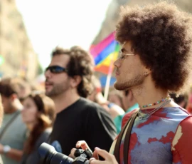 Francoise · 03/10/2026 · 2400 x 1600 Un homme avec une coiffure afro volumineuse, vêtu d’un t-shirt coloré, tient un appareil photo lors d’une manifestation