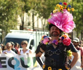 Francoise · 03/10/2026 · 2400 x 1600 Personne participant à une manifestation avec un costume noir décoré de fleurs colorées et un grand chapeau floral, souriant