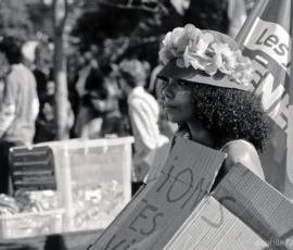 Cyrille · 03/10/2026 · 2026 x 1621 Une personne participant à une manifestation porte un carton avec un message et un chapeau fleuri, entourée d'autres