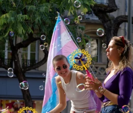 Cyrille · 03/10/2026 · 2026 x 1520 Deux personnes souriantes jouant avec des bulles de savon devant un grand parapluie coloré dans une rue animée.