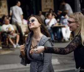 Cyrille · 03/10/2026 · 2026 x 1520 Deux femmes souriantes en mouvement dans un lieu public, l'une portant une veste à carreaux et l'autre une tenue élégante