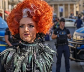 Marc · 03/10/2026 · 1080 x 1080 Personne déguisée avec une perruque orange volumineuse et une tenue fantaisiste en extérieur, entourée de policiers en tenue