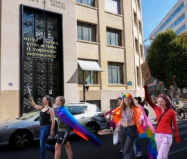 Marc · 03/10/2026 · 1080 x 1080 Groupe de personnes participant à une manifestation devant un bâtiment institutionnel, brandissant des drapeaux arc-en-ciel