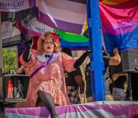 Marc · 03/10/2026 · 1080 x 1080 Personne déguisée en costume extravagant rose et violet sur une scène festive avec des drapeaux arc-en-ciel et des bannières