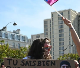 Aurélie · 1287 x 2048 Manifestante tenant une banderole en français avec un drapeau arc-en-ciel, dans une rue urbaine ensoleillée.