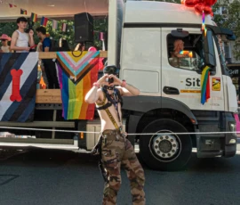 Léonard · 03/10/2026 · 2400 x 2274 Un participant torse nu, vêtu de camouflage, fait un geste avec les mains près d'un camion décoré pour une parade festive