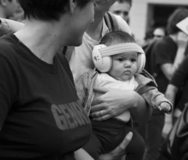 Alice · 03/10/2026 · 1920 x 2400 Un adulte porte un bébé équipé d’un casque audio en milieu urbain, entouré de passants flous en arrière-plan.