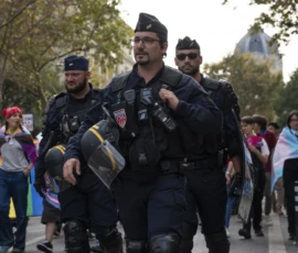 Alice · 03/10/2026 · 2400 x 1600 Deux policiers en tenue anti-émeute marchant au milieu d'une manifestation avec des participants en arrière-plan.