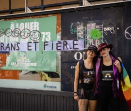 Muriel · 03/10/2026 · 2048 x 1365 Deux personnes souriantes posant devant une affiche et un mur tagués, l’une avec un drapeau arc-en-ciel, dans un quartier