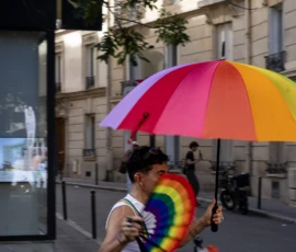 Muriel · 03/10/2026 · 2048 x 1365 Personne marchant en ville sous un parapluie aux couleurs du drapeau LGBTQ+ avec un bracelet arc-en-ciel, ambiance urbaine