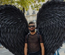 Frédéric · 03/10/2026 · 2400 x 1602 Un individu porte des ailes artificielles noires en plumes et un t-shirt sombre, posant devant un fond urbain avec