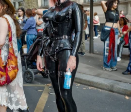 Frédéric · 03/10/2026 · 1121 x 1680 Personne vêtue d'une combinaison noire et brillante avec des accessoires métalliques, marchant en ville parmi un groupe