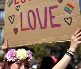 Fabienne · 03/10/2026 · 1603 x 2400 Une personne tenant une banderole colorée avec le message 'Love is Love' lors d'une manifestation, entourée de fleurs et