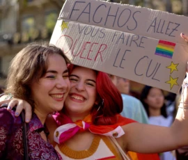Fabienne · 03/10/2026 · 2400 x 1600 Deux personnes souriantes tenant une banderole colorée lors d'une manifestation avec des symboles LGBTQ+ et des étoiles