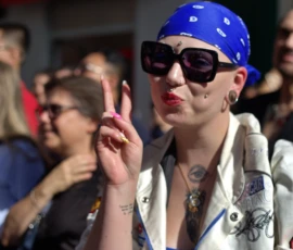 Fabienne · 03/10/2026 · 2400 x 2400 Personne en bandana bleu et lunettes de soleil faisant un geste de la main avec un accessoire rose, vêtue d'une veste