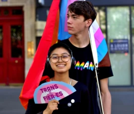 Fabienne · 03/10/2026 · 1600 x 2400 Deux personnes tenant des drapeaux et pancartes LGBTQ+ lors d'une manifestation, ambiance colorée et militante en ville.