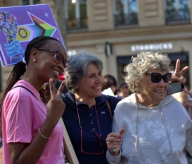 Fabienne · 03/10/2026 · 2400 x 1600 Trois participantes souriantes à une manifestation, l'une tenant une pancarte colorée, toutes faisant des gestes positifs