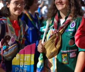 Fabienne · 03/10/2026 · 1282 x 1920 Deux jeunes en tenues de scoutes souriantes participant à une manifestation avec des drapeaux colorés et des accessoires