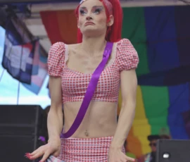 Fabienne · 03/10/2026 · 2400 x 2400 Personne sur scène vêtue d'une tenue à carreaux rouges et d'une ceinture violette, posant avec une expression neutre devant