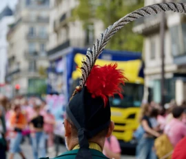 Xavier · 03/10/2026 · 2400 x 1600 Personnage participant à une fête ou défilé portant un chapeau orné de plumes rouges et noires parmi une foule en mouvement.