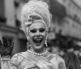 Laurence · 03/10/2026 · 1350 x 900 Personne souriante en costume élégant et perruque volumineuse faisant un geste de la main, ambiance festive et théâtrale