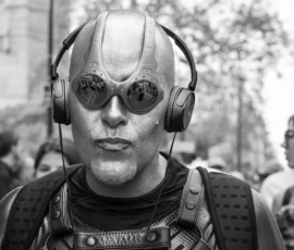 Laurence · 03/10/2026 · 1350 x 900 Personnage en costume futuriste avec casque et lunettes à écrans, portant des écouteurs dans une foule en extérieur