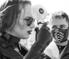 Laurence · 03/10/2026 · 1350 x 900 Une personne en cuir et gants utilise un ventilateur en forme de fleur près d'une autre personne masquée à motifs.